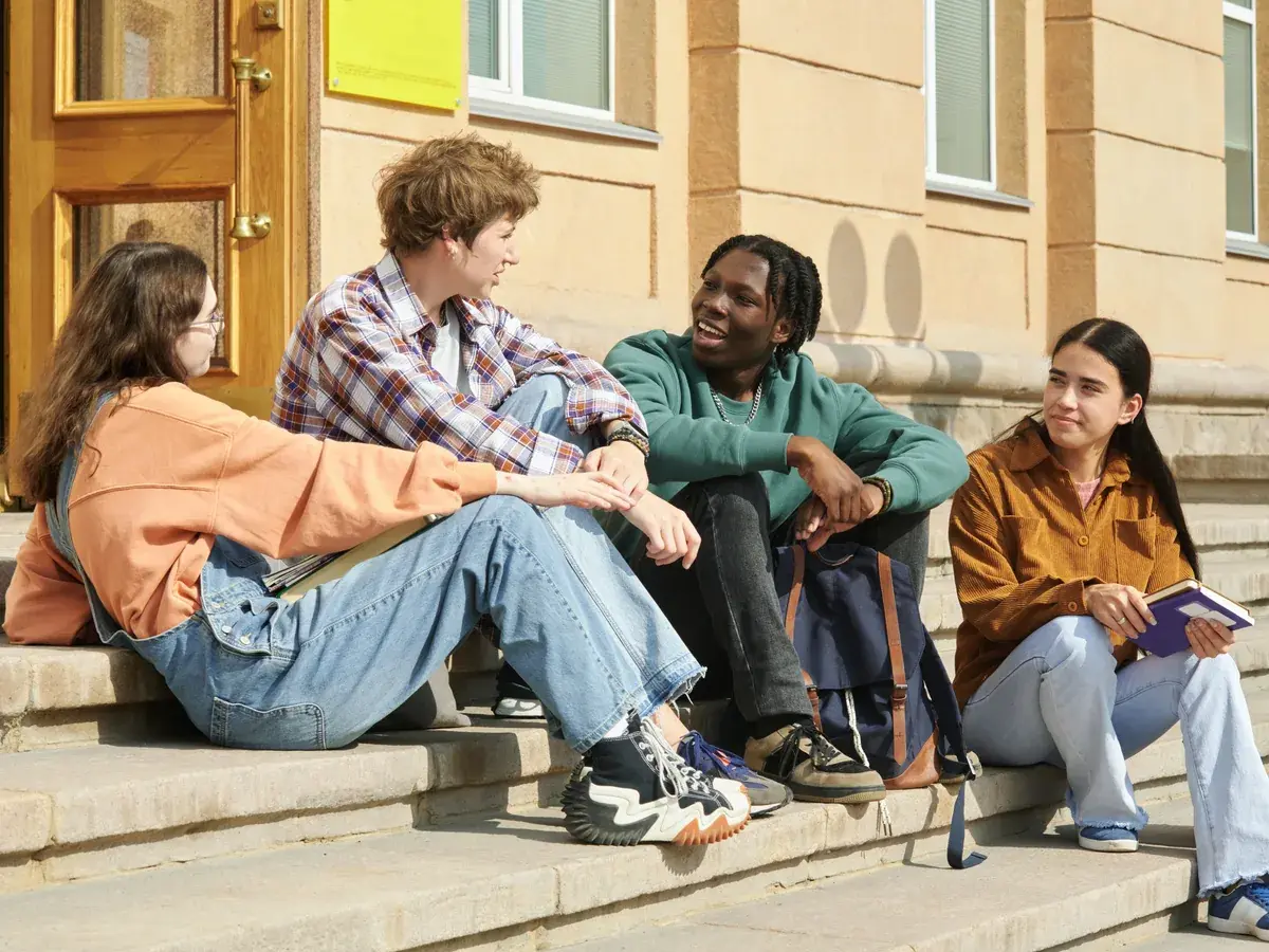 Vier Jugendliche sitzen in der Nachmittagssonne auf den Stufen vor einem Schulgebäude und unterhalten sich